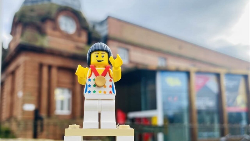 A Lego figure of an athlete wearing a gold medal stands on a podium outdoors. Kelvin Hall can be seen behind the figurine. 