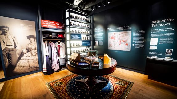 A warmly lit museum exhibit within Mackintosh at the Willow features vintage clothing, numerous tea containers on shelves, a central round table with artefacts, and informative wall displays.