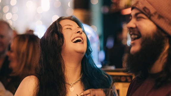  A young woman with long dark hair, a nose ring, and a gold heart necklace laughing heartily, holding a glass, standing next to a bearded man in a beanie.
