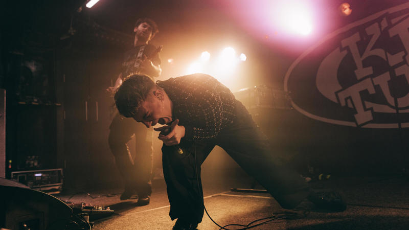 Gemini said A singer in a checked shirt performs passionately on a hazy stage at King Tut's Wah Wah Hut, with a guitarist and bright stage lights in the background.