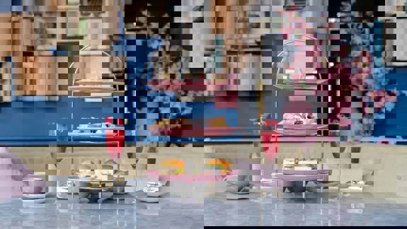 An elegant afternoon tea set on a marble table with a three-tiered stand of sandwiches, scones, and pastries, flanked by pink drinks.