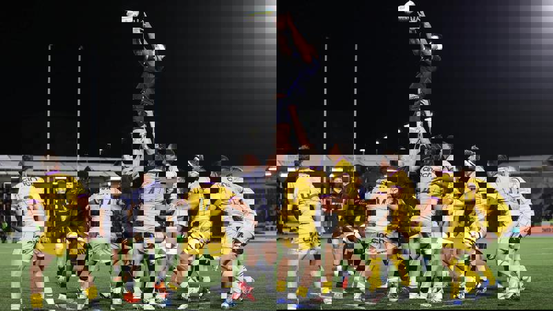 An exciting night-time rugby lineout, with a player leaping high to catch the ball above a cluster of players in yellow and dark jerseys.