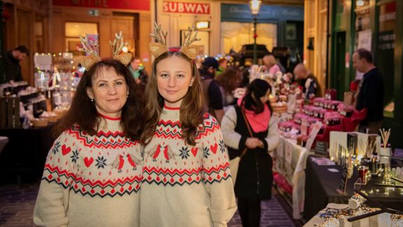 Two people wearing matching festive sweaters with red and white patterns and decorative reindeer antler headbands, standing in front of a market stall filled with holiday-themed items. Other shoppers and stalls are visible in the background.