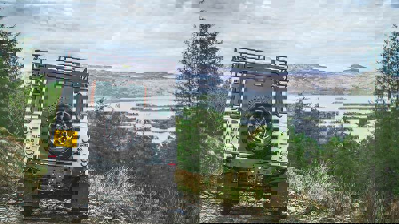 A white Land Rover on a rugged off-road trail overlooking Loch Lomond and surrounding hills.