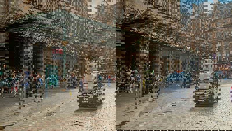 People walk past the historic entrance of Glasgow Central Station, while a black taxi drives along the cobbled street.