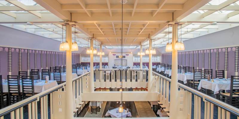 View from the upper level of a large dining room with high ceilings and rows of tables set with white tablecloths. Pendant lights hang from a coffered ceiling, and the space features decorative railings and glass panels.