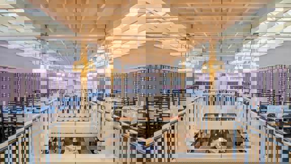 View from the upper level of a large dining room with high ceilings and rows of tables set with white tablecloths. Pendant lights hang from a coffered ceiling, and the space features decorative railings and glass panels.