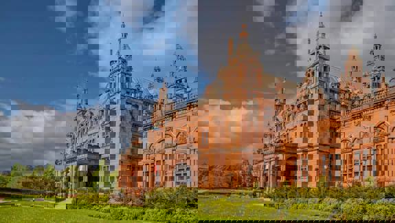 A large, ornate red sandstone building with multiple towers and a curved entrance is surrounded by a green lawn and trees under a blue sky.