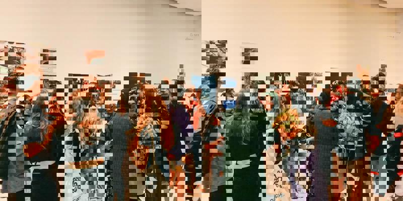 A crowded art gallery opening with a diverse group of people viewing photographs and installations on white walls. In the foreground, a woman holds a bouquet of flowers.