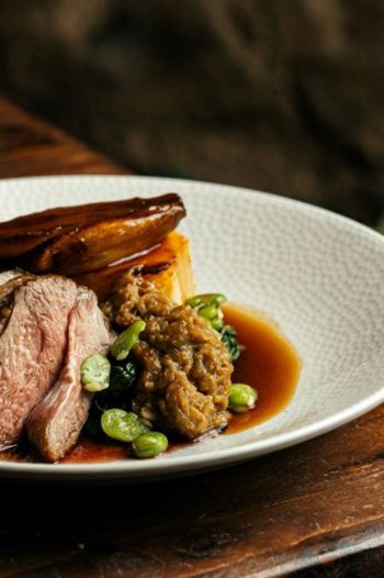 A plate of sliced, medium-rare roast meat (likely venison or lamb) resting on a wooden surface with broad beans and a rich jus.