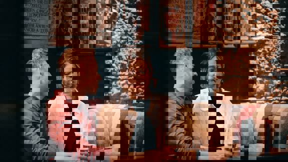 Two people sit close together on a patterned booth in a dimly lit cosy pub. Both are looking contented. In the background, stone walls and framed portraits are visible.