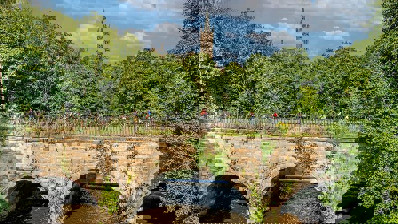 Cyclists crossing a stone bridge with the University of Glasgow tower in the background.