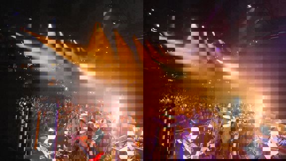 A band on stage from behind, with their arms around each other, facing a large, brightly lit crowd.