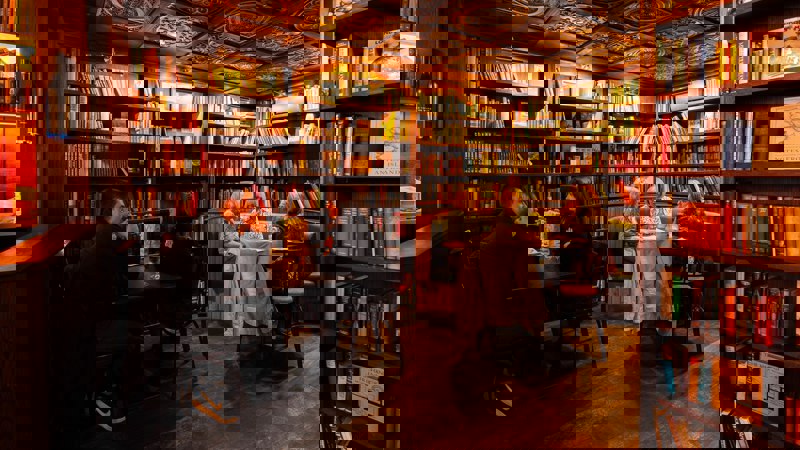 A dark, cosy bar and bookstore with wood-panelled walls and a patterned ceiling. Bookshelves filled with books surround the room. People sit at tables, enjoying drinks.