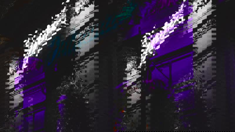 An exterior shot of a building at night, with a bright purple neon sign that reads 'Delmonica's' shining above the entrance.