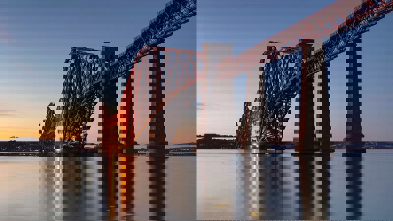 The iconic red Forth Bridge, a cantilever railway bridge, stretches over calm water at dusk, its lights glowing.