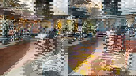 A spacious, modern hotel bar with panoramic riverside views, sleek wooden floors, plush grey armchairs, and ambient lighting with purple and blue hues.