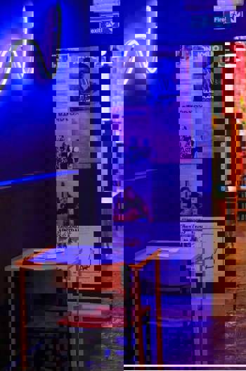 A cozy, dimly lit cafe interior with a small table, a chair, posters on the walls, and a blue neon light.