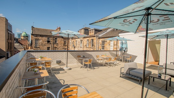 Outdoor rooftop terrace with wooden tables, metal chairs, and large umbrellas, surrounded by brick buildings under a clear blue sky