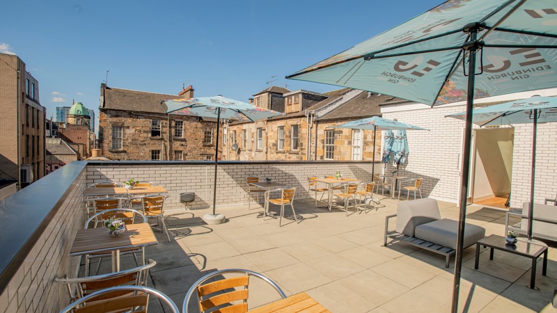 Outdoor rooftop terrace with wooden tables, metal chairs, and large umbrellas, surrounded by brick buildings under a clear blue sky