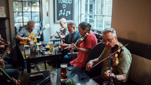 A group of people play traditional music in a pub. The musicians are seated at a table with pints of beer, playing fiddles and a guitar.