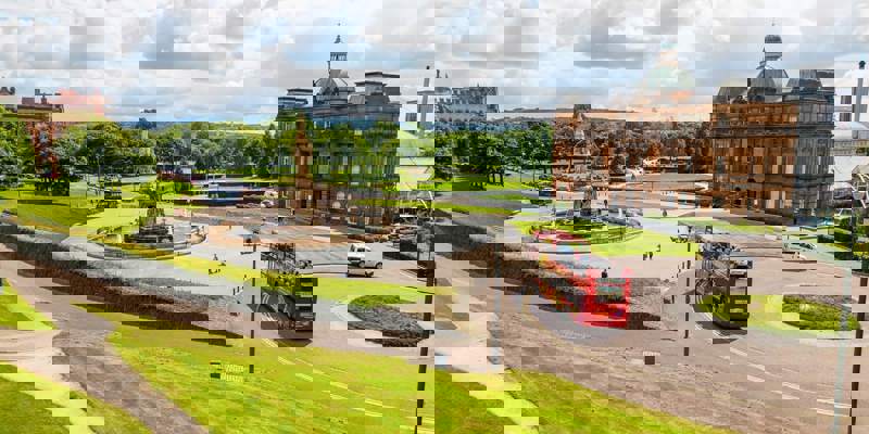 A red double-decker city sightseeing bus driving past the Doulton Fountain and the People's Palace in Glasgow Green on a sunny day.