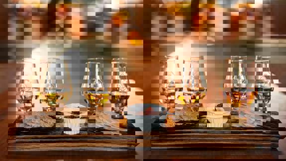 Four glasses of amber whisky are arranged on a wooden serving board with cheese and crackers.