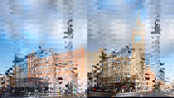 A street-level view of the Tolbooth Steeple at a city intersection, with historic buildings and cars under a cloudy blue sky.