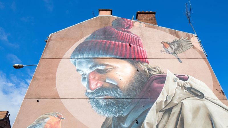 A large, detailed mural of a bearded man in a red hat with a robin on each side of him, painted on the side of a building.