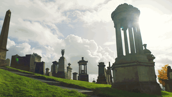 Rows of grand stone monuments and obelisks in a historic cemetery, illuminated by bright sunlight filtering through a dramatic cloudy sky.