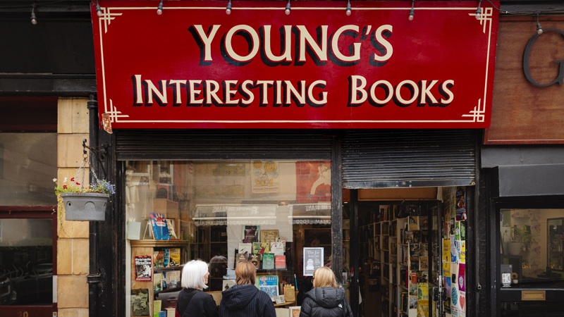 Exterior shot of Young's Interesting Books, a shop with a large red sign and a window display of books. Three people are standing near the entrance, looking inside.