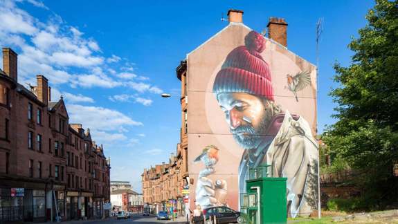 Large street art of a man with a red beanie holding a bird, painted on the side of a building.
