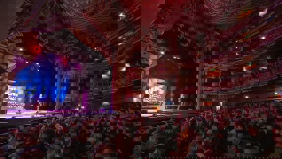 An ornate theater filled with an audience watching a comedian on stage during the Glasgow International Comedy Festival.