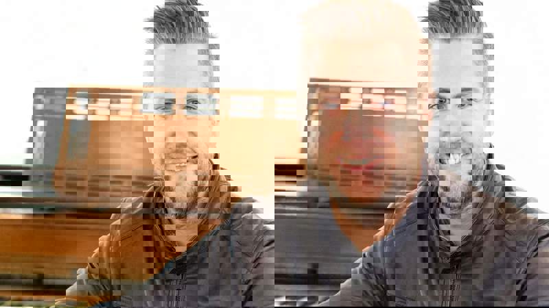 A smiling man in a black leather jacket sitting at a piano with a set of bagpipes on his lap.