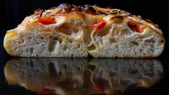 A cross-section of focaccia bread, showing its airy, large-holed interior and a crust topped with tomatoes and rosemary, reflected on a dark surface.