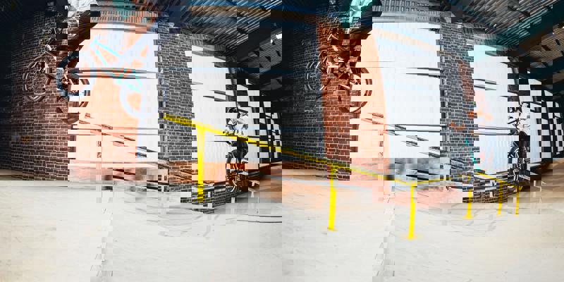 Two bikers on a wooden ramp, one mid-air performing a trick and the other looking on, in an indoor skate park.