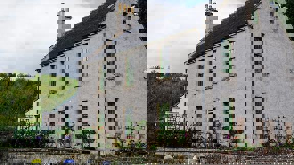 A light-colored, two-story stone building with green-framed windows is surrounded by greenery and a small garden area
