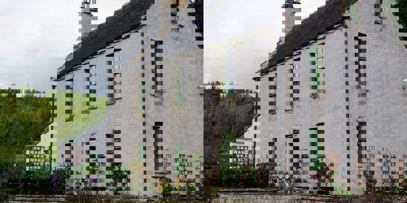 A light-colored, two-story stone building with green-framed windows is surrounded by greenery and a small garden area