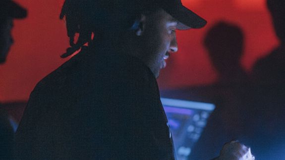 A DJ with dreadlocks and a baseball cap focuses on a mixing console in a dark club. Atmospheric red and blue lights glow in the background.