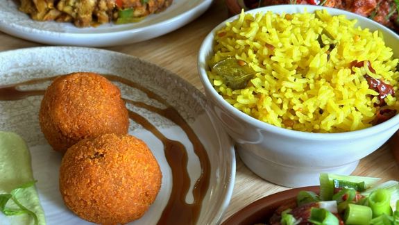 A table filled with various Indian dishes, including yellow rice, fried snacks, a vegetable stir-fry topped with green onions, spicy fried chicken, and a mixed vegetable curry garnished with lemon and chilies