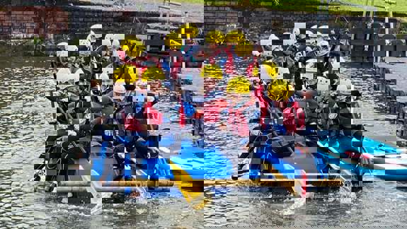A group of children in wetsuits and yellow helmets paddling a raft made of blue barrels on a body of water.