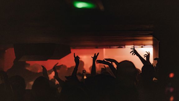 A low-light photo shows a crowd's silhouetted hands raised toward the ceiling under red and green ambient lights in a dark club.