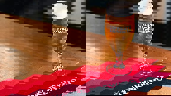 A glass of beer with "Drygate Brewing Co" on it sits on a wooden table with a red, black, and white mat.