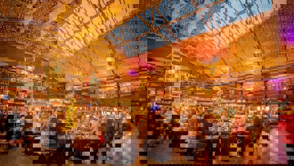 A wide shot of an indoor market with a vaulted glass ceiling, decorated with many small lights and a Christmas tree.