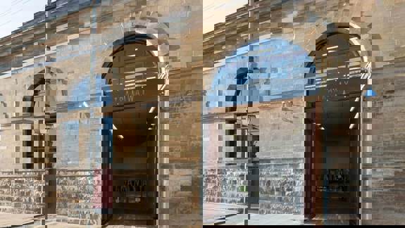 The stone facade of a large building with a wide arched entrance and a smaller arched window to the left. The name 'TRAMWAY' is written above the main entrance.