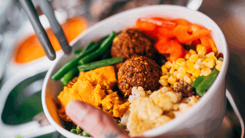 Bowl with green beans, red bell peppers, corn, mashed sweet potatoes, cauliflower, and falafel, held by hand from bottom.