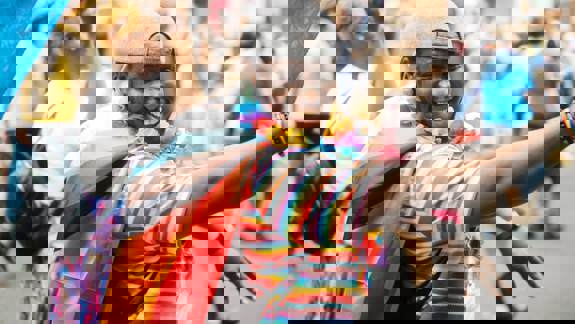 A smiling person wearing a rainbow-striped shirt, a Progress Pride flag like a cape, a lei, and a grey cap celebrates at a Pride event.