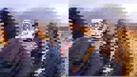 A group of tourists, one in a kilt, stand on a rural road in front of a Rabbie's tour bus with mountains in the background. 