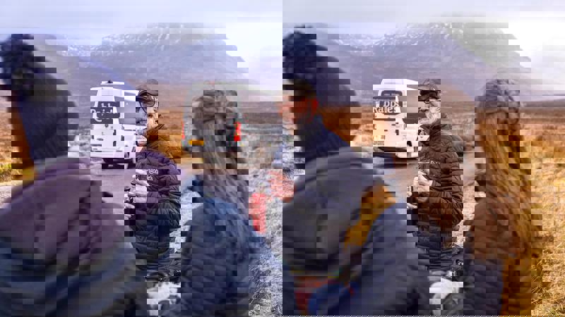 A group of tourists, one in a kilt, stand on a rural road in front of a Rabbie's tour bus with mountains in the background. 