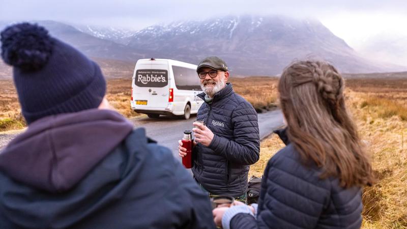 A group of tourists, one in a kilt, stand on a rural road in front of a Rabbie's tour bus with mountains in the background.